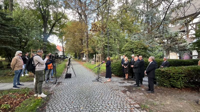 Kobieta przemawia do mediów podczas obchodów 25-lecia wpisu Kościoła Pokoju w Świdnicy na Listę Światowego Dziedzictwa UNESCO. Stoi przy mikrofonach na terenie obok świątyni, a za nią zgromadzona jest grupa uczestników wydarzenia. Po lewej stronie operatorzy kamer i fotografowie rejestrują wystąpienie. W tle widać parkową alejkę, drzewa oraz fragment zabudowy przy Kościele Pokoju.