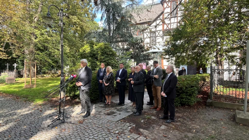 Prof. Jacek Purchla przemawia do mediów podczas obchodów 25-lecia wpisu Kościoła Pokoju w Świdnicy na Listę Światowego Dziedzictwa UNESCO. Stoi przy mikrofonach na brukowanej alejce, a za nim zgromadzona jest grupa uczestników wydarzenia. W tle widoczne są drzewa, zabytkowe nagrobki oraz fragment szachulcowej bryły Kościoła Pokoju.