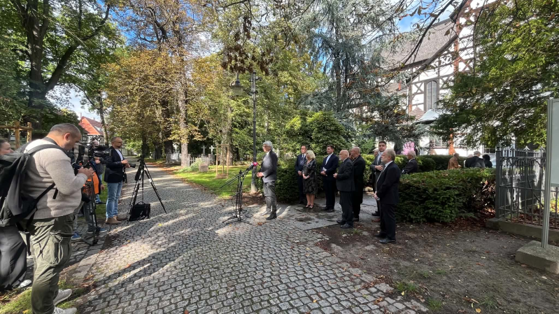 Prof. Jacek Purchla przemawia do mediów podczas obchodów 25-lecia wpisu Kościoła Pokoju w Świdnicy na Listę Światowego Dziedzictwa UNESCO. Stoi przy mikrofonach na alejce przed świątynią, a obok niego zgromadzeni są uczestnicy wydarzenia. Po lewej stronie operatorzy kamer i fotografowie rejestrują wystąpienie. W tle widać drzewa oraz fragment szachulcowej bryły Kościoła Pokoju.