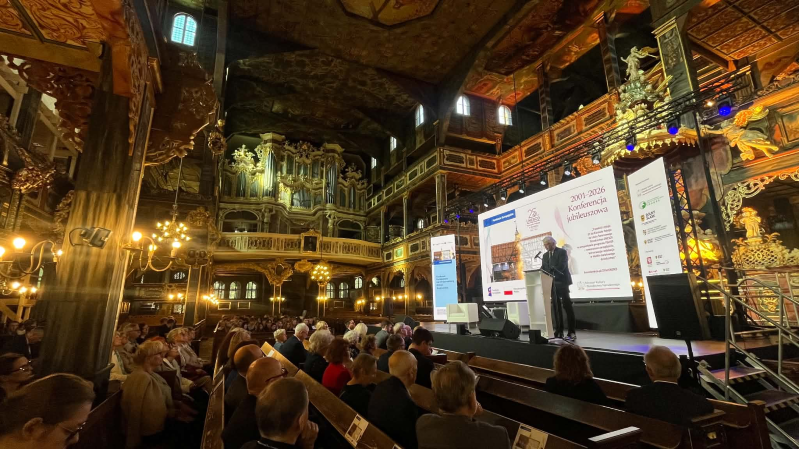Prof. Jacek Purchla przemawia na scenie podczas konferencji z okazji 25-lecia wpisu Kościoła Pokoju w Świdnicy na Listę Światowego Dziedzictwa UNESCO. Wydarzenie odbywa się we wnętrzu bogato zdobionej świątyni; przed sceną siedzi licznie zgromadzona publiczność, a w tle widoczne są zabytkowe organy, empory i dekoracje kościoła.