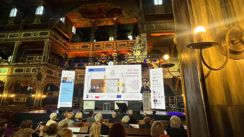 Prof. Jacek Purchla przemawia na scenie podczas konferencji z okazji 25-lecia wpisu Kościoła Pokoju w Świdnicy na Listę Światowego Dziedzictwa UNESCO. Wydarzenie odbywa się we wnętrzu bogato zdobionej świątyni; przed sceną siedzi licznie zgromadzona publiczność, a w tle widoczne są zabytkowe organy, empory i dekoracje kościoła.