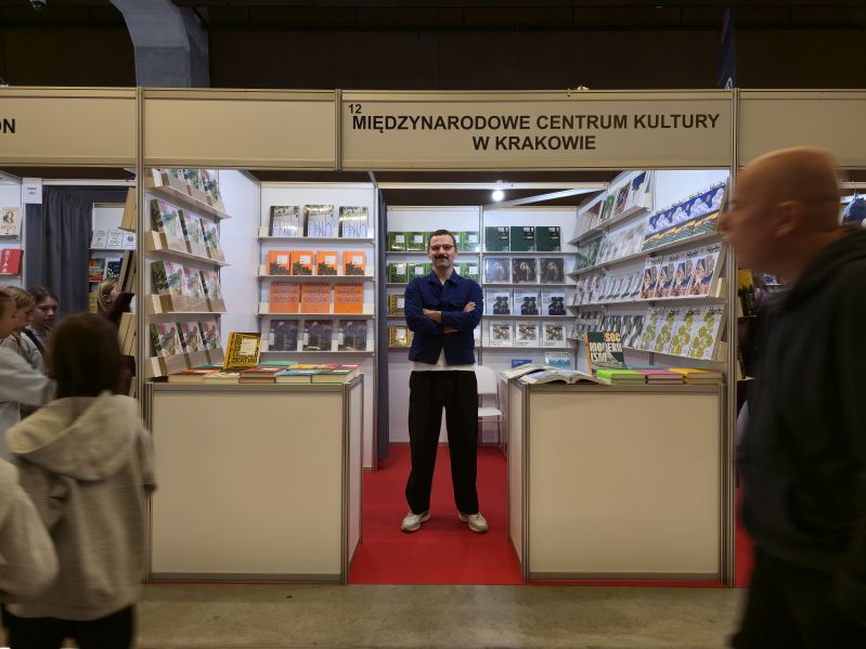 Bartosz Sadulski na stoisku Międzynarodowego Centrum Kultury w Krakowie podczas Gdańskich Targów Książki 2026. Stoi pośród prezentowanych książek wydanych przez MCK i numerów magazynu HERITO, a przed stoiskiem przechodzą odwiedzający targi.