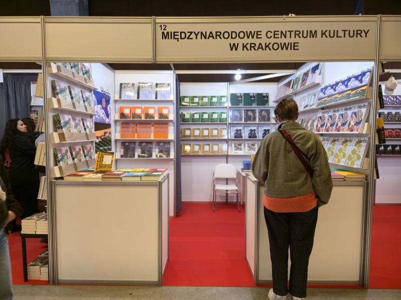 Stoisko Międzynarodowego Centrum Kultury w Krakowie podczas Gdańskich Targów Książki 2026. Odwiedzająca ogląda prezentowane na półkach i ladach książki wydane przez MCK oraz numery magazynu HERITO.