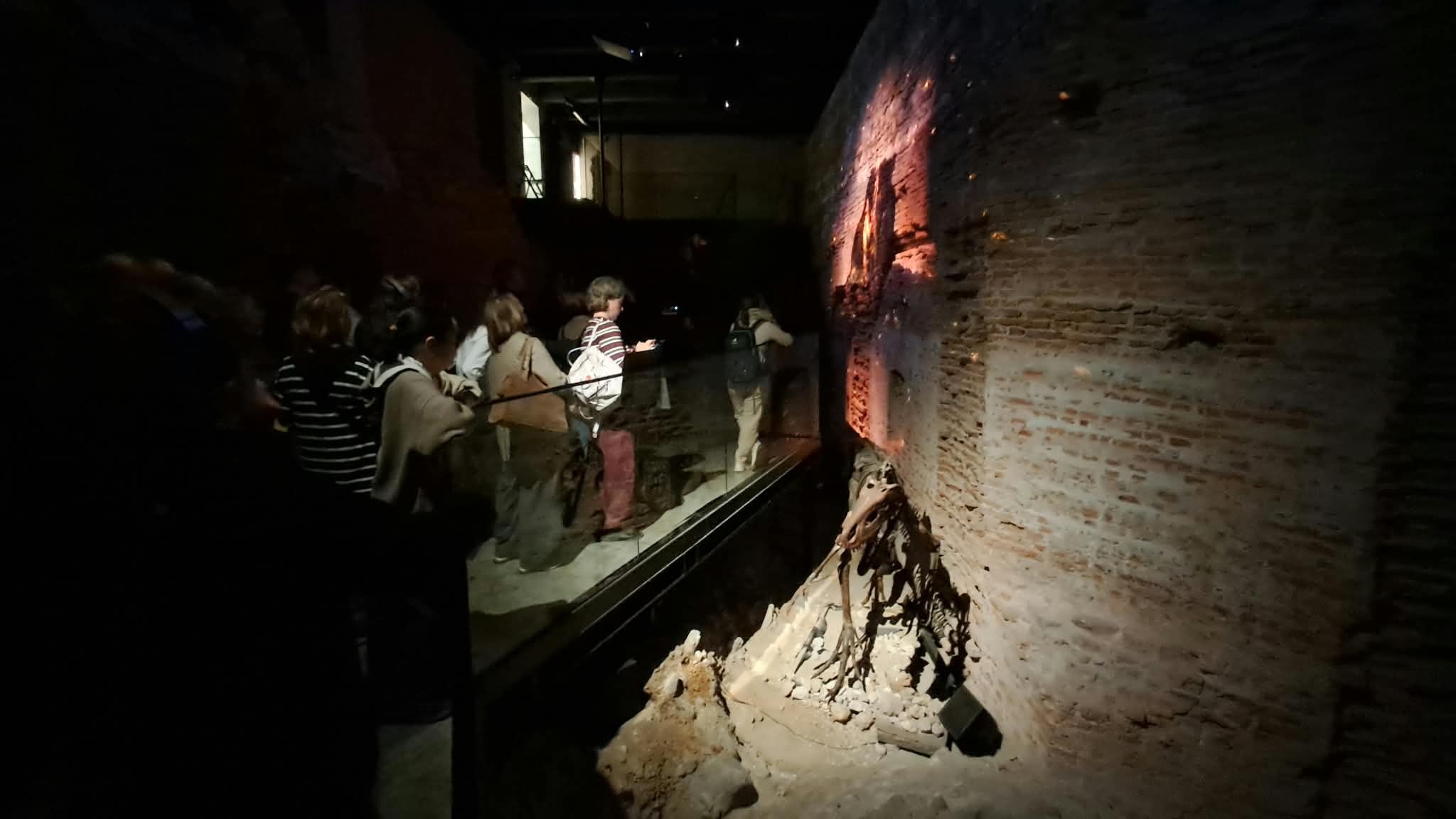 Uczestnicy zwiedzają podziemną trasę ekspozycyjną na Wawelu. Przechodzą wzdłuż zachowanych ceglanych murów i reliktów archeologicznych, oglądanych w przyciemnionym, punktowo oświetlonym wnętrzu.