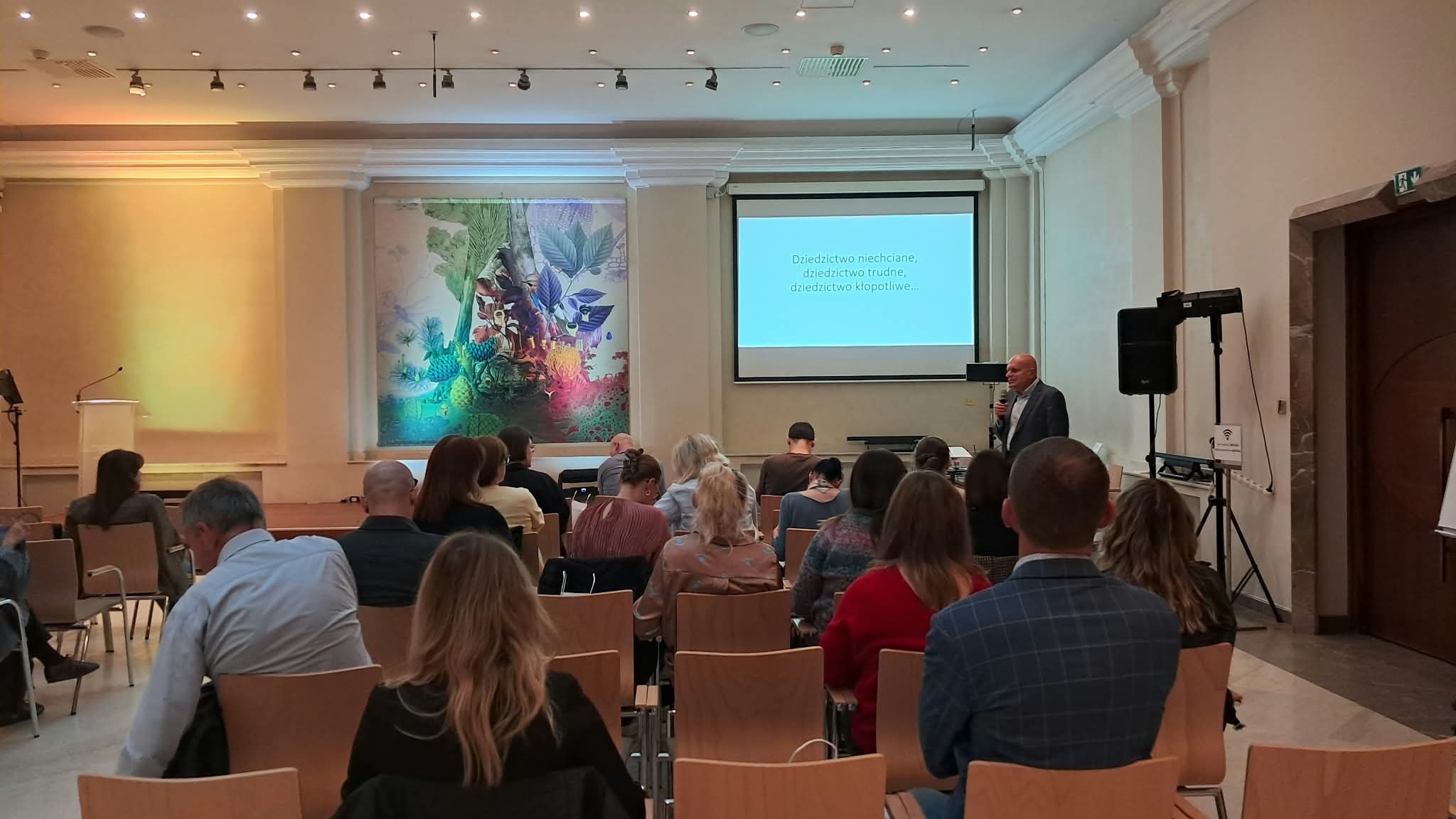 Prelegent z mikrofonem prowadzi wykład dla uczestników spotkania w sali Pod Kruki MCK. Na ekranie wyświetlony jest slajd z hasłem Dziedzictwo niechciane, dziedzictwo trudne, dziedzictwo kłopotliwe, a publiczność siedzi w rzędach i słucha wystąpienia.