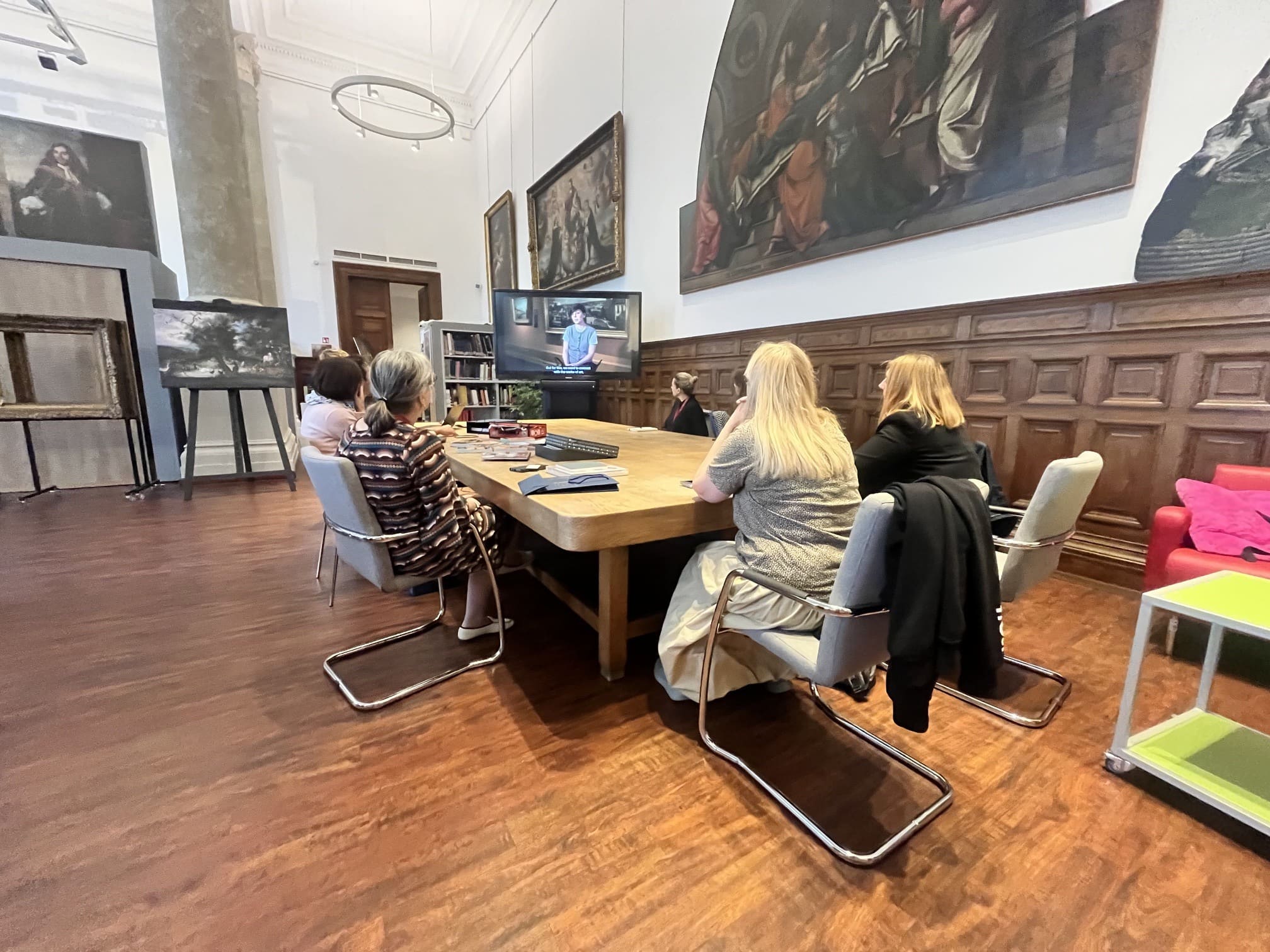 Przedstawicielki Międzynarodowego Centrum Kultury podczas wizyty studyjnej w jednej z budapeszteńskich instytucji muzealnych. Grupa siedzi przy dużym stole i ogląda materiał wideo wyświetlany na ekranie; wnętrze zdobią obrazy, drewniana boazeria i regały z książkami.