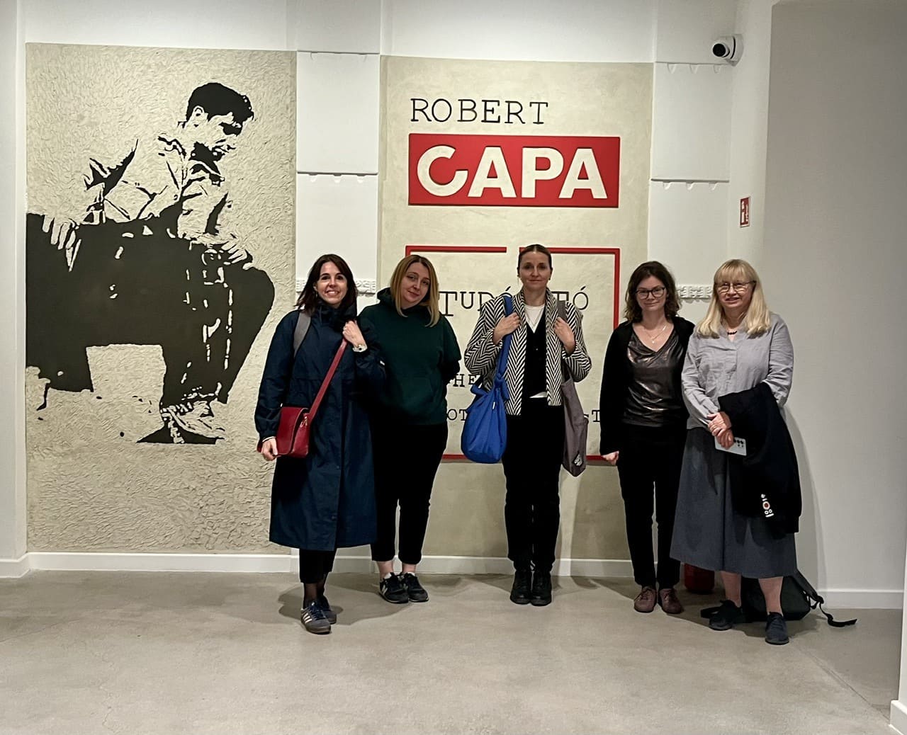 Pięć przedstawicielek Międzynarodowego Centrum Kultury podczas wizyty studyjnej w Centrum Fotografii Współczesnej im. Roberta Capy w Budapeszcie. Kobiety pozują do wspólnego zdjęcia przed ścianą z dużym napisem ROBERT CAPA i graficznym wizerunkiem fotografa.