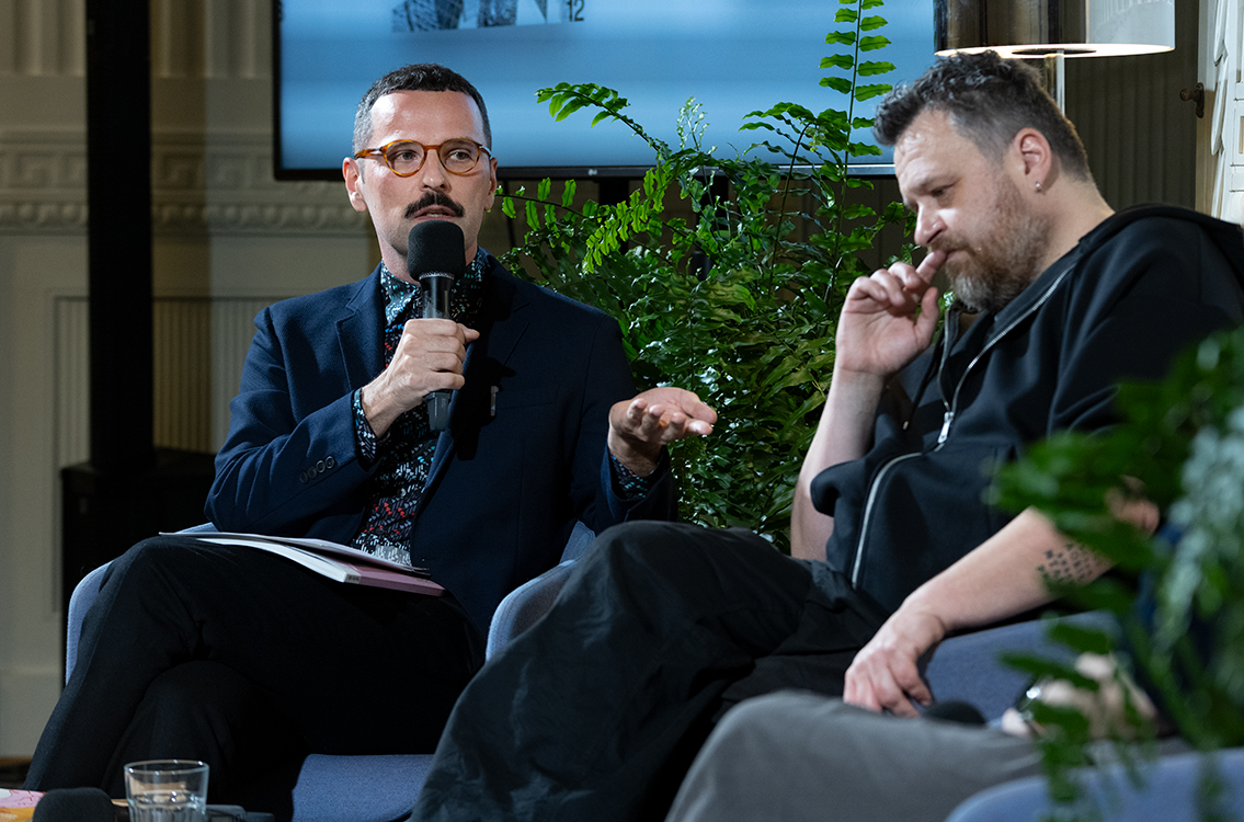 Bartosz Sadulski przemawia do mikrofonu podczas spotkania poświęconego 60. numerowi magazynu HERITO, gestykulując w trakcie wypowiedzi. Obok niego siedzi Ziemowit Szczerek, słuchający rozmowy.