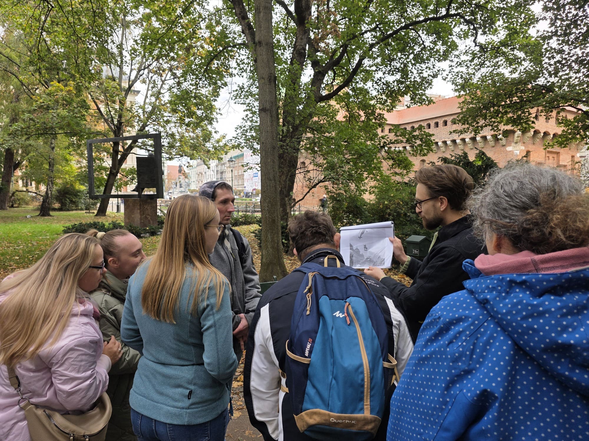 Marcin Krotla prowadzi spacer po Plantach w ramach Festiwalu Kultury bez Barier dla osób z Teatru Drużyna. Uczestnicy stoją wokół prowadzącego i oglądają pokazywaną przez niego historyczną ilustrację; w tle widoczny jest fragment Barbakanu i zieleń Plant.