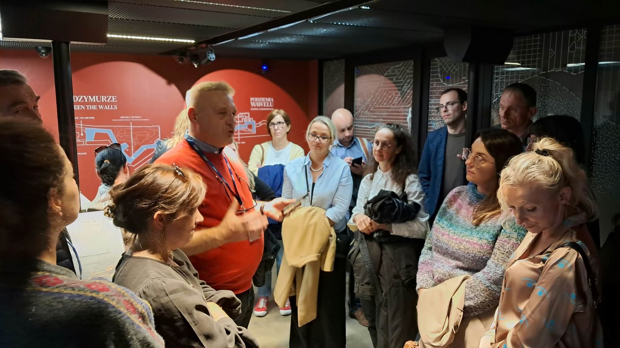 Przewodnik oprowadza grupę uczestników po ekspozycji w podziemiach Wawelu, objaśniając prezentowane zabytki i historię miejsca. Zwiedzający stoją wokół niego w niewielkiej sali, a w tle widoczne są plansze informacyjne dotyczące podziemi i dawnych murów zamkowych.