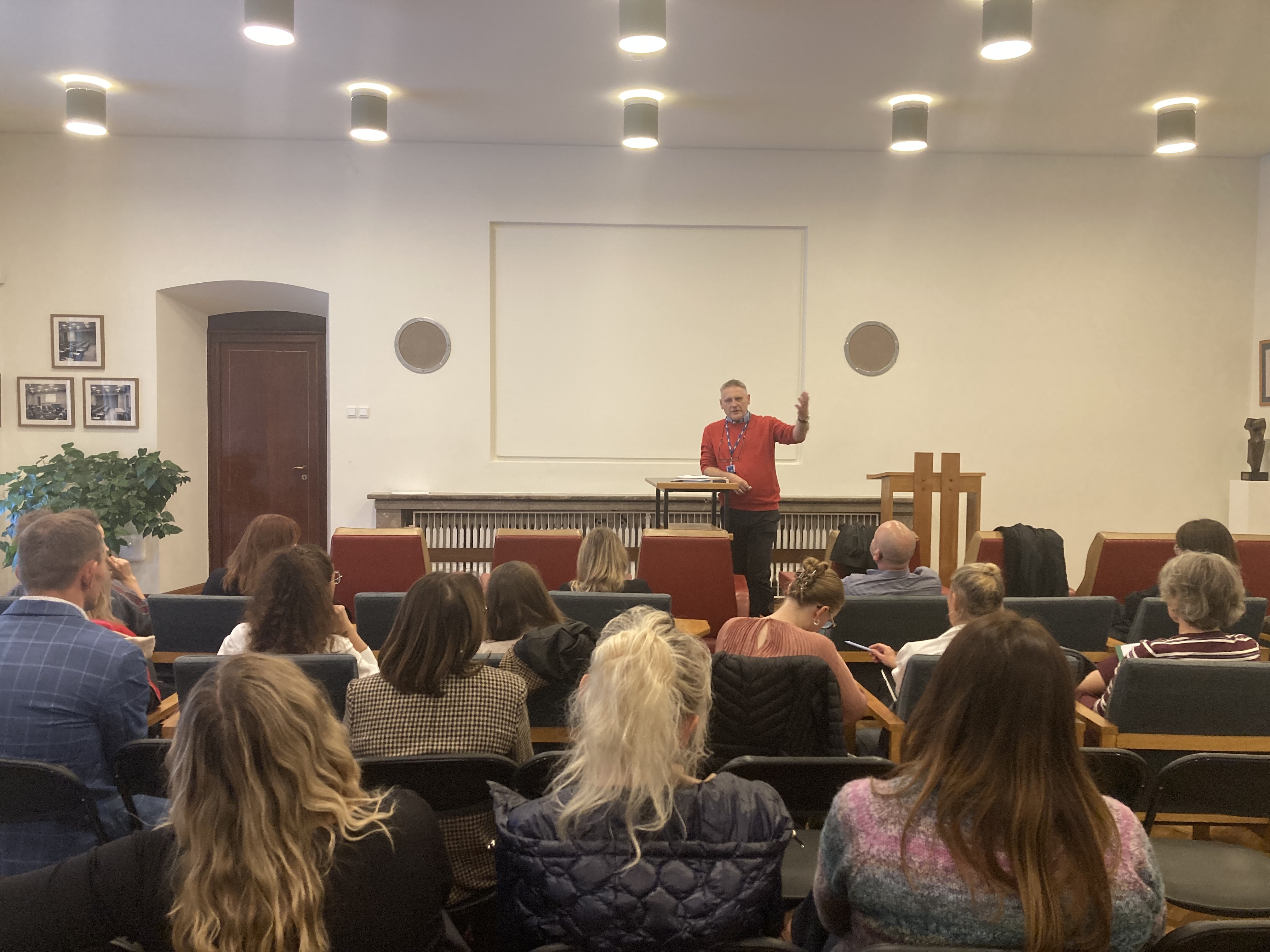 Mężczyzna prowadzi prelekcję dla grupy uczestników siedzących w sali na Wawelu. Stoi przy pulpicie, gestykulując podczas wystąpienia, a słuchacze zajmują miejsca w rzędach foteli.