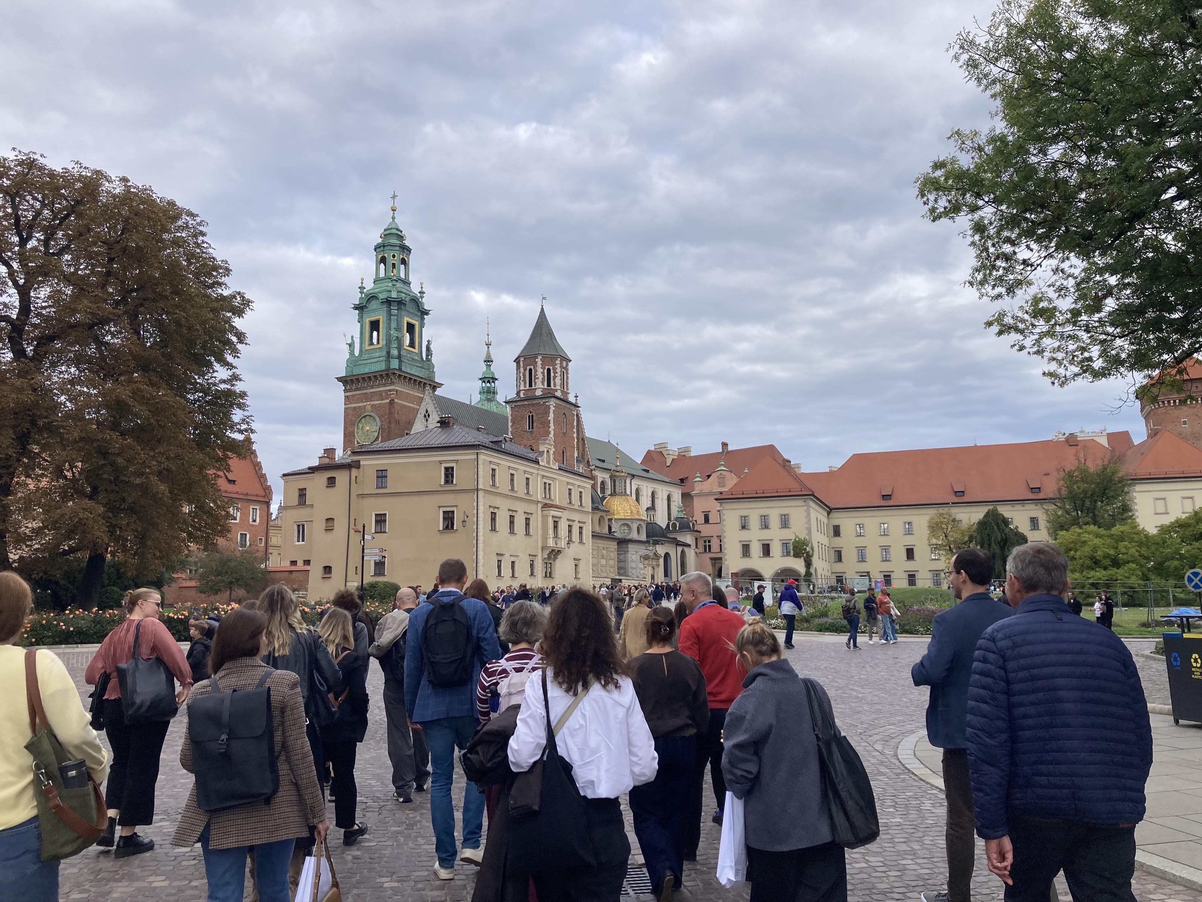 Grupa uczestników spaceruje w kierunku zabudowań Wawelu w Krakowie. W tle widoczne są wieże katedry wawelskiej, zabytkowe budynki zamkowe oraz dziedziniec otoczony zielenią.